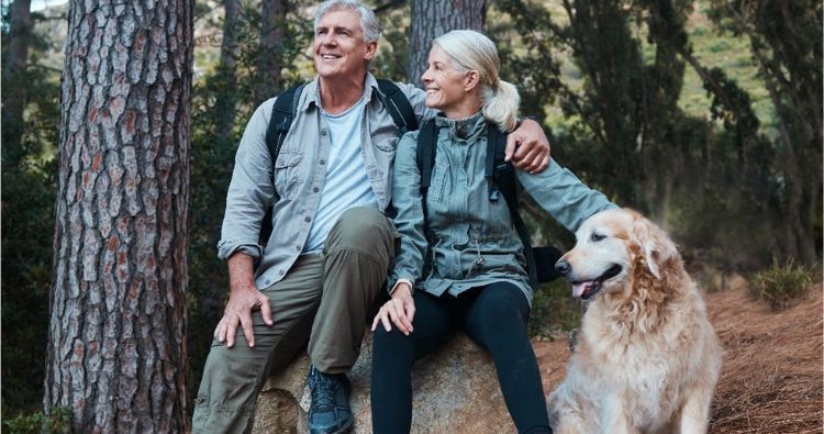 An older couple sit in the woods with their dog.